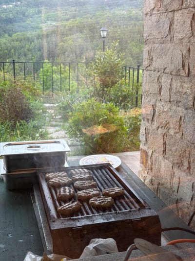 The image shows a grill with patties being cooked, possibly burgers or similar meat products. Smoke is rising from the patties. Next to the grill is a metal container and some plastic bags. The scene seems to be outdoors, with a view through a window or screen, showcasing a landscape of trees and what appears to be a lamp post in the distance. There is a fence or railing visible amidst the greenery. A stone wall is visible in the upper portion of the image. The location is Кирково, Bulgaria. There are no people visible in the image.