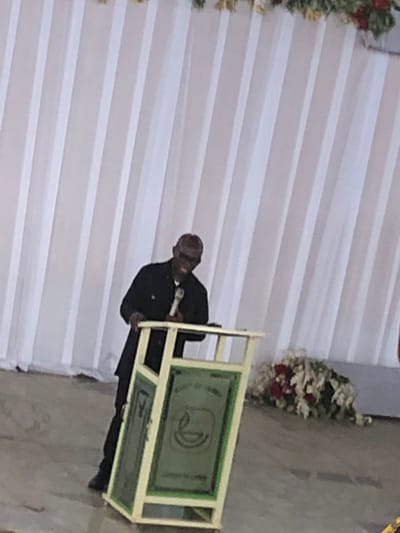 A man in a dark suit stands at a podium with a microphone. The podium is cream-colored and decorated with green panels displaying a symbol. Behind the man, a white pleated curtain hangs and is decorated with floral arrangements. The location is Nteje, Nigeria.