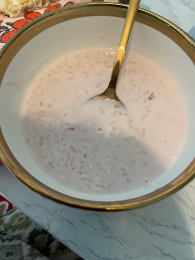 A close-up shot shows a bowl of rice pudding with a golden spoon in it. The bowl has a golden rim and is on a marble-patterned table, partially covered by a colorful patterned cloth. In the upper left corner, a piece of bread with butter is visible. The background is blurred. The scene appears to be indoors.