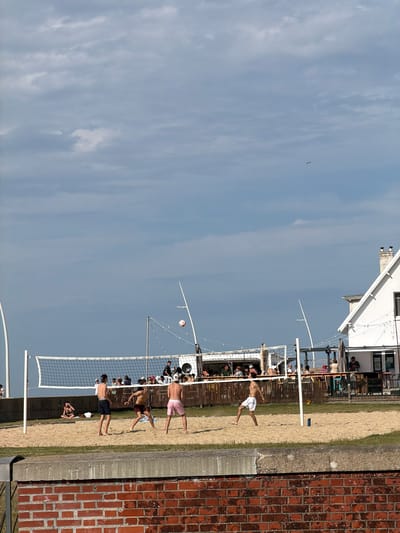 The image depicts a beach volleyball game at Hautot-sur-Mer, France. Several people are actively playing or spectating the game on a sandy court next to a building. The game appears to be in progress, with players in various positions and one player mid-air, suggesting an active moment in the game. The setting is outdoors, with a clear sky and some clouds visible. The time of day appears to be daytime, likely afternoon, given the lighting and shadows. A volleyball net is set up, and a volleyball is visible in the air. The building in the background has a white facade and appears to be a structure associated with the beach, possibly a cafe or changing rooms. A brick wall is visible on the right side of the image, providing a textured contrast to the sandy environment. The overall atmosphere seems casual and recreational, with people enjoying a sporting activity on a pleasant day.