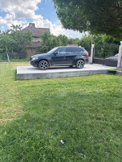 The scene depicts a black SUV parked on a concrete driveway adjacent to a green lawn. The driveway leads to a black metal gate. In the background, a building with a tiled roof is visible, partially obscured by trees. The sky above is blue with scattered clouds. The photograph was taken in Septemvritsi, Bulgaria.