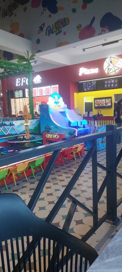 The image captures an indoor entertainment area, likely a children's play zone or amusement arcade, situated in Hrazdan, Armenia. The foreground is dominated by a black metal railing, partially obscuring a colorful floor patterned with circles in shades of cream, green, and brown. In the mid-ground, a bright blue and purple slide with a cartoonish top is a prominent feature, surrounded by what appears to be an interactive game or ride. Several brightly colored chairs—red, yellow, and green—are neatly arranged along a counter or waiting area, illuminated by a strip of multicolored lights.
Behind the play structure, a red wall displays a prominent "Pizza" sign in glowing white lettering, accompanied by a circular graphic depicting a slice of pizza. Adjacent to this, a yellow tiled wall features a framed menu with images of food items like tacos and fries. Further in the background, a sign that reads "EFC" is visible, along with what looks like a fast-food restaurant menu. The ceiling is decorated with a playful mural featuring colorful shapes and the words "MANCHO LAND" and a rocket graphic. A few individuals are present, including a person seated behind the counter and others in the distance, suggesting the establishment is operational. The overall atmosphere appears lively and geared towards family entertainment.