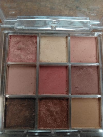The image displays a clear plastic eyeshadow palette containing nine square pans of cosmetic product, situated on a light-colored, textured surface in Ciudad de San José, Costa Rica. The pans are arranged in a 3x3 grid.
The top row, from left to right, contains:
1. A shimmery rose-gold eyeshadow, exhibiting minor depressions and texture variations indicative of previous use.
2. A matte cream-colored eyeshadow, with a smooth, largely undisturbed surface.
3. A matte light peach-pink eyeshadow, with a smooth, largely undisturbed surface.
The middle row, from left to right, contains:
4. A matte light brown eyeshadow, with a smooth, largely undisturbed surface.
5. A matte medium rose eyeshadow, with a smooth, largely undisturbed surface.
6. A matte reddish-brown eyeshadow, with a smooth, largely undisturbed surface.
The bottom row, from left to right, contains:
7. A shimmery dark brown eyeshadow, with a largely undisturbed, textured surface.
8. A shimmery warm brown/copper eyeshadow, with a largely undisturbed, textured surface.
9. A matte light tan eyeshadow, with a smooth, largely undisturbed surface.
The clear plastic hinged lid of the palette is open and partially visible at the top of the frame, showing slight reflections and minor surface scratches. The underlying surface, likely a tabletop, is out of focus. No individuals, actions, or interactions are present in the scene.