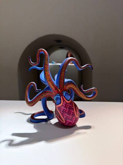 An ornate, wooden octopus sculpture is mounted on a white wall. The octopus has blue and brown striped tentacles, a deep red body with a geometric pattern, and one visible eye. The sculpture casts a shadow on the wall behind it. To the left, a circular recessed feature is visible in a gray wall, creating a contrasting element in the background. The lighting is soft, accentuating the details of the sculpture and its shadow.