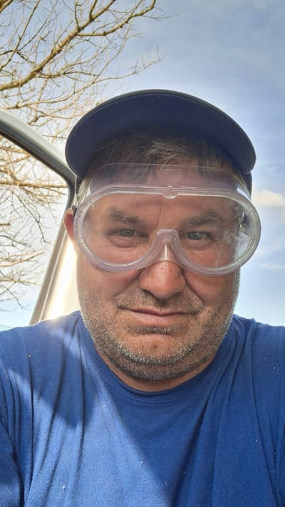 This media file is a close-up, rotated selfie of a middle-aged man wearing a navy blue baseball cap and clear safety goggles. He has a stubbly beard and is wearing a navy blue t-shirt. The man is looking directly at the camera, with a neutral expression. The background shows a bright, partly cloudy sky and the bare branches of trees, suggesting an outdoor setting in a temperate climate, possibly during autumn or winter. The image appears to have been taken during the daytime, with natural light illuminating the scene. There are small specks of what appears to be sawdust or debris visible on the man's t-shirt and around the safety goggles. The perspective suggests the man might be in or near a vehicle, indicated by a sliver of a car frame visible in the lower left of the frame. The setting is Hîncești, Moldova. No text is visible in the image.