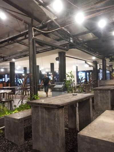 The image displays an indoor or semi-open-air commercial dining space within an establishment in Pontianak, Indonesia. The ceiling features an exposed industrial aesthetic with black metal trusses, large black pipes, and multiple bright, circular overhead light fixtures. The floor is covered with dark grey gravel. Seating consists of numerous tables and accompanying benches constructed from unpolished concrete, each topped with a dark wooden slab.
In the mid-ground, a male individual, wearing a dark short-sleeved shirt and dark shorts, walks barefoot from the right towards the left side of the frame. To his right, a light grey lidded trash bin is visible. Various green and red-hued potted plants are interspersed throughout the seating area, defining sections. On the wooden surface of the large concrete table in the foreground, a small white ashtray is present. White electrical conduits are attached to the concrete supports of some tables. Multiple dark-colored structural pillars are spaced across the area. In the background, additional tables and chairs are visible, with several indistinct figures seated, suggesting active patronage. The scene is evenly illuminated by the overhead lighting.