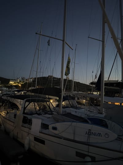 The image captures a marina scene at twilight or early night within the Lavreotiki Municipal Unit, Greece. Numerous sailboats are moored, occupying the foreground and midground, with their masts and rigging extending into a deep blue-grey sky. A small Greek flag is visible flying from the rigging of a sailboat towards the upper left portion of the image. Another sailboat displays a colorful flag, patterned with yellow, green, and blue, from its mast more centrally located.

In the immediate foreground, a sailboat's hull is visible, with two paddleboards strapped securely to its starboard side; the prominent one clearly displays the brand "WINDY" along with a circular logo. Several boats exhibit deployed dark fabric covers over their cockpits. Illuminated interiors of boat cabins and various dock lights cast warm glows throughout the marina. Text indicating "southseasail.com" is legible on the boom of one vessel, and "White Blue Seas" is visible on the hull of another boat further back.

The background features a dark, undulating hillside upon which several buildings are situated. These structures, some appearing residential and others possibly under construction, are illuminated by internal and external lights. Additional streetlights or public lighting sources are visible on the hillside and within the marina. No individuals are present or engaged in observable actions within the frame.