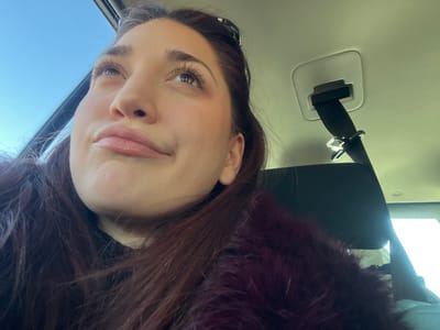 An indoor image, set within a vehicle in Dorlisheim, France, captures a young adult female from a low-angle perspective. The subject occupies the left side of the frame, looking upward and slightly to her right, with a subtly pursed lip expression. She has long, dark reddish-brown hair, with sunglasses resting on top of her head. She is wearing a dark garment under a prominent, textured burgundy faux fur coat. To the right, a segment of the vehicle's light-colored headliner is visible, from which a black seatbelt extends downward, attached to a chrome-finished buckle mechanism. A black car seat headrest is also partially visible on the right. The background outside the vehicle shows a clear blue sky, indicating daylight conditions.