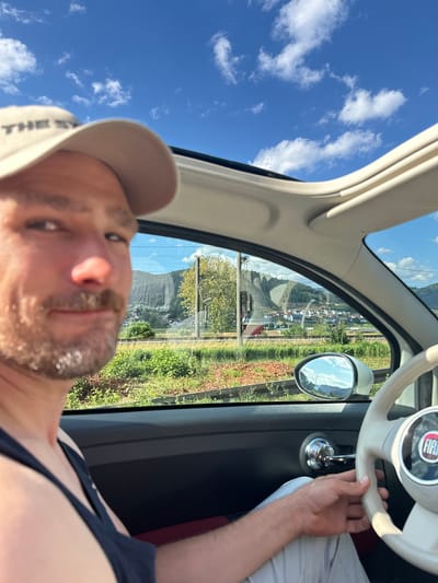 In the city of Haslach im Kinzigtal, Germany, a man wearing a beige baseball cap and a dark tank top is inside a white Fiat 500 car, holding the steering wheel. The car's sunroof is open, revealing a bright blue sky with scattered white clouds. The man is looking towards the camera, with a mustache and goatee. Through the car windows, a scenic view of a town nestled in a valley with rolling green hills and trees is visible. A train track runs horizontally across the landscape. The side mirror reflects the sky and distant mountains. The interior of the car shows a cream-colored dashboard and steering wheel with the Fiat logo.
