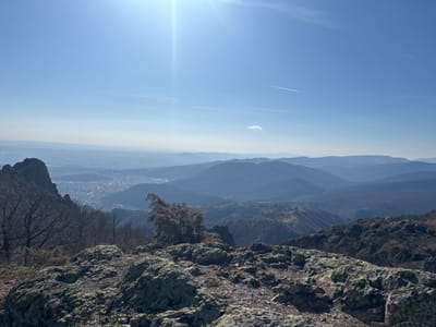 The image displays a panoramic mountain landscape overlooking a city under a clear blue sky. The vantage point is identified as a mountain within or bordering the Sinite Kamani Nature Park, near the village of Ichera, Bulgaria, providing a view of the city of Sliven in the distant valley.
In the immediate foreground, rugged rock formations covered with pale green and grey lichen dominate the lower portion of the frame. Sparse, bare deciduous trees are visible on the left, suggesting a dormant season. The midground features a sprawling urban area, the city of Sliven, characterized by a dense concentration of buildings within a valley, bordered by undulating hills. Beyond the city, a series of progressively lighter blue mountain ranges extend into the hazy distance, demonstrating atmospheric perspective. The sky occupies the upper half of the image, presenting a vivid blue expanse. A prominent sun glare emanates from the upper left quadrant. A faint white aircraft contrail is visible diagonally across the upper right sky, accompanied by a small, isolated white cloud. No human figures or discernible actions are present.