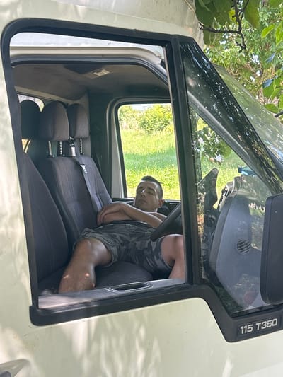 This is a medium shot, taken from outside a white truck cab, looking through an open door and a tilted windshield. The scene depicts a young man, appearing to be in his late teens or early twenties, sleeping slumped in the passenger seat of the truck. He is wearing camouflage shorts and a grey t-shirt. The truck's interior is visible, with dark fabric seats and headrests. The windshield reflects the surrounding environment, showing green foliage and trees, suggesting an outdoor setting in daylight, possibly during warm weather. Outside the truck, parts of tree branches and leaves are visible in the upper left corner, casting shadows on the windshield. The overall impression is one of rest or a break in a journey. Text visible on the exterior of the truck, near the windshield, reads "115 T350". The location context is Lovets, Bulgaria.