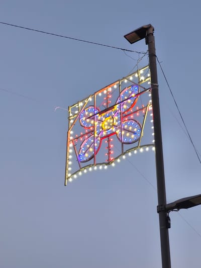 The image shows a street lamp in San Salvo, Italy, with a decorative light display attached to it. The light display is rectangular and is illuminated with a variety of colored lights, including white, red, and blue. The display features a floral or butterfly-like pattern in the center, with smaller lights forming the outline of the rectangle. The sky is a pale blue, indicating it is either early morning or late evening. There are no people visible in the image. The street lamp itself is gray and metallic, with a modern design. There are also thin wires extending across the frame, likely power lines or cables. The overall scene is festive and suggests a public space decorated for an event or holiday.