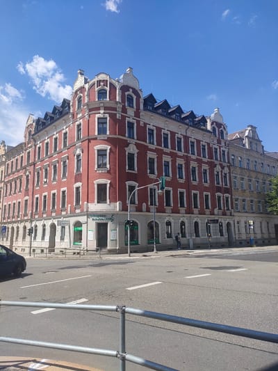 A street-level, eye-level, full shot captures the corner of a red brick building with white ornate detailing and mansard roofs in Chemnitz, Germany. The building is a multi-story structure with numerous windows, some of which are arched. On the ground floor, there are several businesses, including "Orthopädie-Schuhtechnik" (Orthopedic Shoetech). A street with traffic lights and road markings is in the foreground. A dark blue car is visible on the left side of the image, and a pedestrian is walking on the sidewalk in front of the building. The sky above is blue with scattered white clouds. Adjacent to the red building is a yellow building with similar architectural features.
