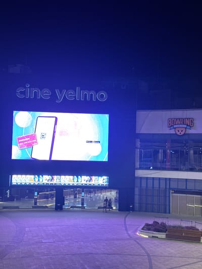 The image shows an outdoor nighttime scene in Alcorcón, Spain. The main subjects are a large digital screen displaying advertisements and a building with illuminated signs. On the digital screen, the text "cine yelmo" is prominently displayed in large white letters, along with an advertisement featuring a smartphone displaying a QR code and a credit card. The screen also shows icons for app stores.
Above the digital screen, on the building facade, a sign for "BOWLING" is visible, with a cartoon bowling ball icon and smaller text below that reads "TRES HORAS". The building itself appears to be a modern commercial establishment with large windows reflecting the lights.
In the foreground, a paved walkway with subtle circular markings can be seen. To the right, there is a planter box with greenery and a wooden bench, illuminated by soft lighting. The overall impression is that of a vibrant entertainment complex at night. Several people are visible in the background near the entrance of the building, suggesting it is open and active. The ambient lighting casts a purple hue over the scene, typical of nighttime urban environments.