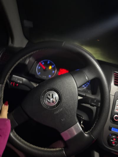 The image depicts the interior of a Volkswagen vehicle at night. A human left hand, adorned with red nail polish and partially clad in a magenta long-sleeved garment, is positioned on the top-left portion of the black, textured steering wheel, which features a central Volkswagen emblem and "AIRBAG" inscription. A grey and white stitched cover is present on the upper left rim of the steering wheel. The dashboard instrument cluster is illuminated, with a tachometer indicating approximately 2000 RPM and a yellow warning light, consistent with a transmission or engine malfunction indicator, illuminated. Additional red warning lights are active on the left side of the dashboard. To the right, a climate control display shows "22.0". Control stalks are visible behind the steering wheel. The exterior visible through the windshield is dark, with a beam of light projecting forward, suggesting headlights are active in an unlit environment. No external visual cues are present to identify a specific location within the city of Batoshevo, Bulgaria.