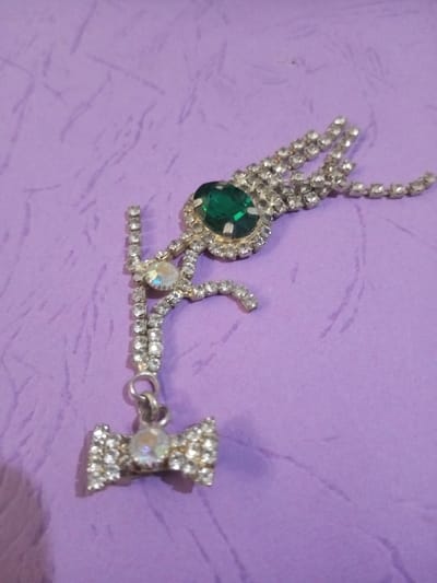 The image displays a single piece of silver-toned costume jewelry, heavily encrusted with numerous clear rhinestones, presented against a light lavender-colored surface that exhibits a subtle, irregular texture. The jewelry item features a prominent oval, faceted emerald-green stone, approximately 1.5 cm in length, positioned centrally towards the upper right, encircled by a halo of smaller clear rhinestones. Multiple short strands of clear rhinestones extend from the green stone towards the upper right. A curved, stylized element, also encrusted with clear rhinestones, extends from beneath the green stone towards the lower left. This element incorporates a larger, iridescent round rhinestone midway along its length. Attached to the lower end of this stylized element by a small silver-toned jump ring is a bow-shaped dangle, approximately 2 cm in width, which is also covered in clear rhinestones and features a central, larger, iridescent round rhinestone. No individuals or specific actions are depicted. The scene is located in the city of Chandragiri, Nepal.