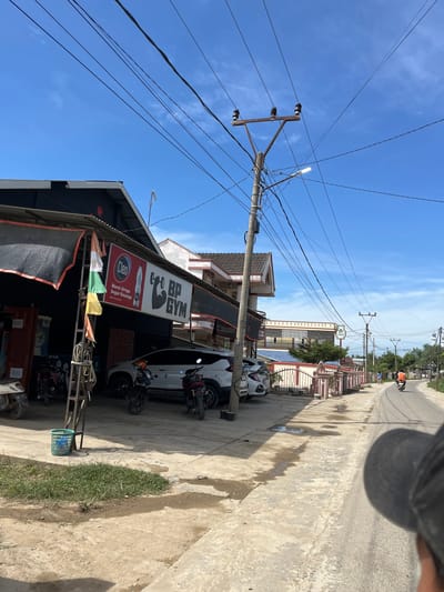A bright, sunny day in a city in Indonesia, where several buildings line a paved road. The buildings have dark roofs, and one of them has a banner that reads "BP GYM". In front of the gym, there are several motorcycles parked. The road itself is wet in places, suggesting recent rain. A few people are visible, including a person riding a motorcycle in the distance. The sky is a clear, vibrant blue with a few wispy clouds. Overhead, a network of power lines crisscrosses the sky, supported by wooden poles. A street lamp is also visible, illuminating the area with a bright light.