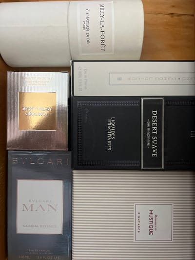 The image displays an overhead view of several perfume boxes arranged on a wooden surface. From top to bottom and left to right, the items are: a tall, white cylindrical bottle with a label reading "CHRISTIAN DIOR PARIS" and "MILLY-LA-FORET"; a rectangular black box with a white label and text that is partially obscured but includes "EAU DE PARFUM 100ML 3.3 FL. OZ."; a silver box with a gold label displaying "TOM FORD METALLIQUE EAU DE PARFUM VAPORISATEUR SPRAY 1.7 FL. OZ./50 ML"; a black box with white text reading "LIQUIDES IMAGINAIRES" and a smaller label with "DESERT SUAVE - EAU imaginaire -"; a dark gray box with the "BVLGARI" logo, a white label with "BVLGARI MAN GLACIAL ESSENCE", and smaller text indicating "EAU DE PARFUM VAPORISATEUR NATURAL SPRAY 100 ML 3.4 FL.OZ. U.S."; and finally, a box with a striped pattern featuring a white label with the text "Mémories de MUSTIQUE" and "EIGHT & BOB". The overall scene is static, with no people or actions depicted. The environment appears to be an indoor setting, possibly a drawer or shelf, due to the wooden texture. There is no information in the image to suggest the location is in Saburovo, Russia.