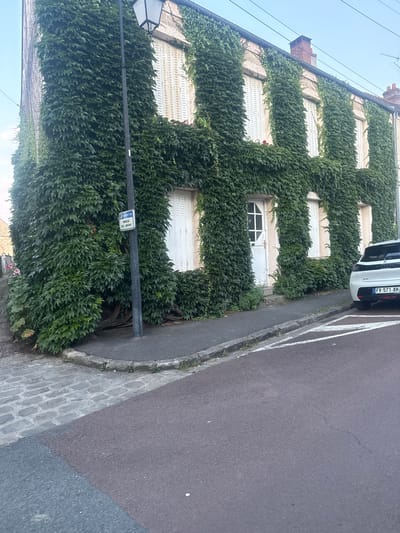 The image shows a corner building in Longpont-sur-Orge, France, extensively covered in ivy. A street lamp stands in front of the building, next to a sign that reads "IMPASSE DES JARDINS". The building has several shuttered windows and a white door. A white car is parked on the street in front of the building, with a license plate that reads "FV-571-AM". The street in front of the building is paved with asphalt, and there is a cobblestone sidewalk on the left side of the image. The sky is clear and blue.