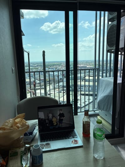 A high-angle medium shot captures a cluttered desk next to a large window overlooking a cityscape and sky. The desk is covered with various objects, including a silver laptop displaying a video of people, a bottle of iced water with a green cap, a bottle of what appears to be a sauce or condiment with a red cap, a white plastic container with a blue label, and a crumpled plastic bag. Several crumpled wrappers and receipts are also visible.
The window reveals a bright, cloudy sky and a dense urban landscape with numerous buildings and what appears to be a railway track. Balcony railings are visible in front of the window, suggesting the viewpoint is from a high-rise building. An air conditioning unit is mounted on the exterior wall. The overall impression is one of a lived-in, somewhat disorganized personal space with a view of a busy city. The lighting suggests it is daytime. The scene is likely set in Sao Thong Hin Town Municipality, Thailand, based on the provided location context.