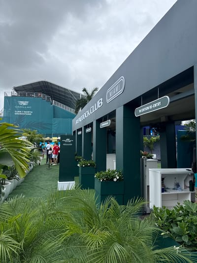 The image shows the Paddock Club entrance at the Miami International Autodrome in Miami Gardens, Florida. The sky is overcast. A large grey awning with the "Paddock Club" logo hangs over the entrance. Signs indicate "Miami" and "W Edition Re-Entry." People are seen queuing at the entrance. A large structure for the Cadillac Turn 1 North Grandstand is in the background. Lush greenery including palms is in the foreground.