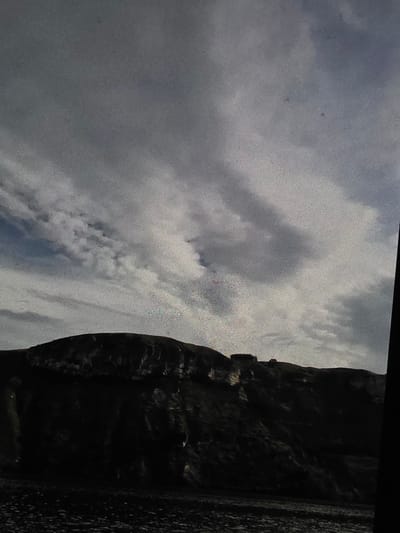 The image depicts a dark, mountainous landscape under a cloudy sky. The foreground is a dark, rocky terrain, possibly a beach or a scree slope. Rising behind it is a steep, dark mountain or cliff face, casting deep shadows. The summit of the mountain is silhouetted against the sky and appears to have some structures on it, possibly buildings or ruins. The sky is filled with dramatic, heavy clouds, with patches of lighter sky visible through them. There are no people or animals visible in the image. The overall impression is one of a dramatic and somewhat bleak natural landscape.
Regarding the location in the city of Dubno, Ukraine, there is no visual information in the image that can definitively place it within Dubno or any specific location. The image shows a generic mountainous or hilly coastal environment.