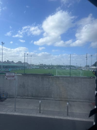 The image shows a view of a sports field in Bray, Ireland, likely a football (soccer) pitch, with a grandstand and surrounding buildings. In the foreground, a concrete wall runs horizontally across the image, with a sidewalk and road in front of it. A metal pole with multiple directional signs is visible to the left of the concrete wall. The signs point towards "MERMAID THEATRE", "TOURIST OFFICE", and "BRAY CENTRE", indicating this is a public area within Bray. The sports field is green and appears well-maintained, with goals set up at either end. Several people, likely players or staff, are scattered across the field. Stadium lights are mounted on poles around the perimeter of the field. The sky is blue with scattered white clouds. The overall scene is typical of a sports facility in a suburban or town setting.