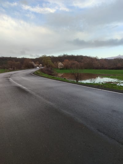 A wet, black asphalt road curves through a rural landscape. A flooded field is visible adjacent to the road, with trees and vegetation surrounding the field. The sky is overcast with gray and white clouds. A line of trees stretches along the horizon. The scene appears to be in a rural or countryside location, and there is no definitive information to confirm this to be Gabar, Bulgaria.