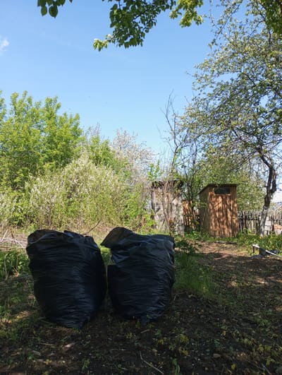 The image depicts two large, black plastic garbage bags situated on a patch of dirt and grass in what appears to be a backyard or garden in Монастырка, Russia. The bags are positioned in the lower center of the frame. Behind the bags, the scene includes various vegetation: bushes with white flowers, green trees, and scattered weeds. Two small wooden structures resembling sheds or outhouses are visible in the middle ground, partially obscured by the foliage. A wooden fence is also discernible in the background. The sky is blue, with some tree branches and leaves visible at the top of the frame. The scene is bathed in sunlight, suggesting it is daytime.