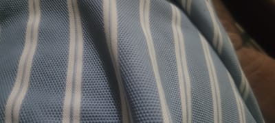 This image is a close-up, macro shot of a blue and white striped fabric. The fabric appears to be textured, possibly a knit or pique material, with a distinct pattern of vertical stripes. The lighting creates shadows and highlights that emphasize the texture and folds of the fabric, giving it a three-dimensional quality. There are no discernible elements in the image that would indicate a location in Kipala Mpakani, Tanzania, or any other specific geographic setting. The subject is solely the fabric itself.