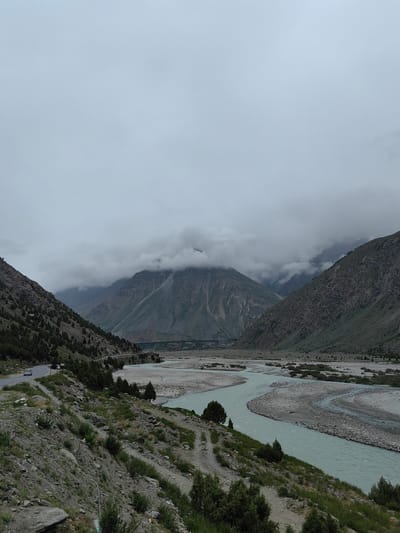The image depicts a wide, meandering river flowing through a valley in Jispa, India. The river has light blue, opaque water and is bordered by a wide, stony riverbed. On the left side of the valley, a paved road curves along the mountainside, with a white car visible driving on it. The slopes of the mountains are covered in sparse vegetation, including shrubs and small trees, with rocky outcrops. The valley floor in the distance appears to be a plain with patches of vegetation and smaller water channels. The upper parts of the mountains are obscured by thick, low-lying clouds, creating a dramatic and somewhat moody atmosphere. The sky is overcast and a muted gray.