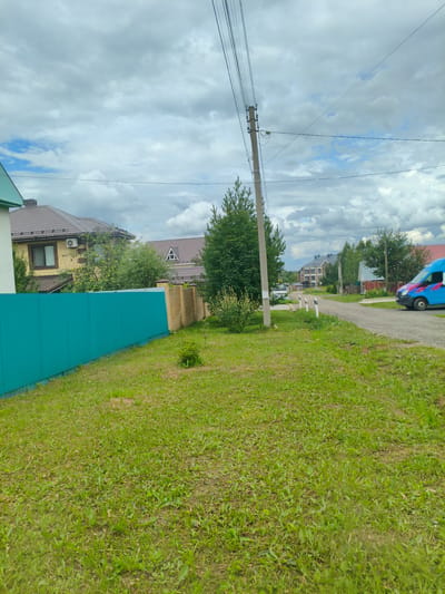 The image depicts a street scene in the village of Khokhryaki, Russia, on a cloudy day. In the foreground, a patch of mowed grass occupies the lower half of the frame, with a bright turquoise metal fence running along the left edge. To the right of the fence, a brick fence is visible. Behind the fences, a two-story house with a brown roof and yellow siding is seen, along with trees. Further down the street, a gravel road curves to the right, lined with more houses and trees. A utility pole with multiple wires stands prominently near the left side of the road. A light-colored van with blue and pink graphics is parked on the right side of the road, facing away from the viewer. The sky is filled with heavy, gray clouds.