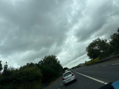 The image shows a grey, cloudy sky over a rural highway in or near Slieverue, Ireland. A silver Volkswagen Polo is in the foreground, traveling on the right side of the road. The car has a license plate reading "D1-CH-64787". In the background, other vehicles are visible on the highway, and trees and greenery line the side of the road. Several houses are also visible in the distance. The overall scene suggests overcast weather conditions.