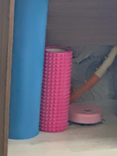The image shows a shelf with several fitness items. On the left is a large, upright blue foam roller. To its right is a pink textured foam roller. Further to the right and slightly behind is an object with a white cylindrical top and a pink cylindrical base, possibly a cleaning tool or another type of roller. In the foreground on the right is a small, round, pink object with legs, appearing to be a miniature robot vacuum or similar device. The background is a textured, grey surface. The overall scene is indoors, likely a storage area or closet. The location of Vladimiro-Alexandrovskoye, Russia is not depicted in the image.