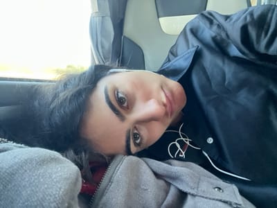 A female individual is depicted lying on her side inside a passenger vehicle, presumably a bus, looking directly into the camera. She has dark, wavy hair, prominent dark eyebrows, and is wearing a dark, collared shirt or jacket with a white snap button visible. White wired earphones are connected, with one earpiece in her right ear and the cable resting on her chest. A grey zippered garment, possibly a jacket or blanket, covers her lower chest and shoulder, partially obscuring a red garment underneath. The background shows a light and dark grey two-tone seat with a headrest, featuring a faint "Intra" logo. To the upper left, a window reveals a bright, blurred exterior with suggestions of green foliage, indicating daylight. The scene suggests travel, consistent with being within or traversing the area of Santarém, Portugal, though no specific city landmarks are visually identifiable.