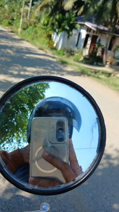 The image is a reflection in a motorcycle's side mirror, taken in Matangbaroh, Indonesia. The mirror shows a person holding a smartphone, wearing a helmet, and an orange sweater. In the background, a road is visible with a house and vegetation. The overall lighting suggests it's daytime.