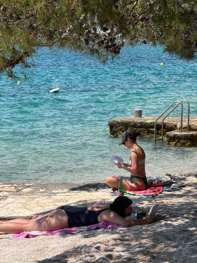 The image shows two women at a rocky beach in Grad Šibenik, Croatia. The woman on the left is wearing a green bikini and a baseball cap, and she is sitting in the shallow water, reading a book. She appears to be tanned and has her hair pulled back. To her right, the woman on the left is lying on a towel on the rocky shore, wearing a black swimsuit. She is also tanned and has dark hair. The water is clear and turquoise, and there are some shadows cast by trees on the shore. The time of day appears to be sunny and warm, suitable for a day at the beach. A metal ladder is visible, leading into the water from the rocky shore. There are some pine trees on the left side of the image.