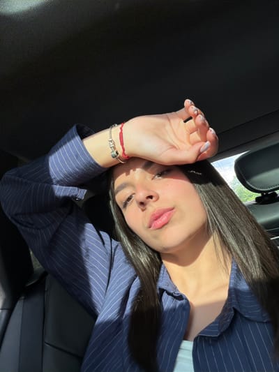 The image shows a woman lying in what appears to be the backseat of a car. The car seat and side panel are visible. The woman is wearing a striped blue shirt. She has long, dark hair. Her hand is raised near her face, possibly shielding her eyes from the sun. The location is not discernible from the image. Хаджийско, Bulgaria is not deducible from the image.