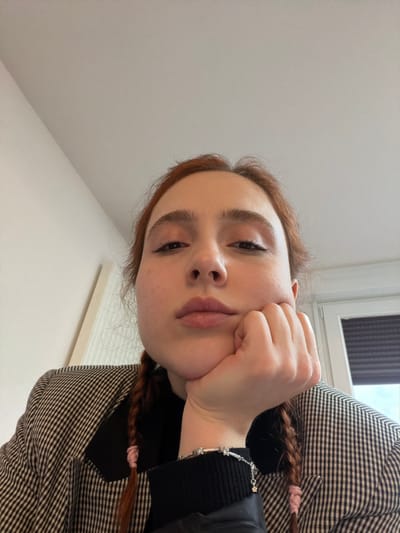 An individual is depicted indoors in Eschau, France, captured from a low-angle perspective. The subject is a person with fair skin and auburn hair styled into two visible braids, one secured with a light pink elastic. They are positioned with their chin resting on their right hand, fingers loosely curled. Their gaze is directed upward towards the camera, and their lips are slightly pursed.
The subject wears a dark-colored, possibly black, turtleneck or high-collared garment underneath a patterned blazer. The blazer exhibits a black and white or dark gray and light gray check or houndstooth pattern. A silver-toned bracelet with multiple small charms, including a star-shaped pendant, is visible on their right wrist.
The background consists of a plain white ceiling occupying the upper portion of the frame. To the left, a light-colored wall is visible, while to the right, a window or door frame with a dark blind or shutter is present, suggesting an exterior view beyond. The lighting is diffuse and even.