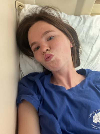 A young woman with brown hair and fair skin, wearing a blue t-shirt, is lying on a white bed with a pillow. She is looking at the camera and puckering her lips. The room appears to be a hospital room, as suggested by the medical equipment visible on the wall and the general sterile appearance. The image was taken in the city of Urman, Russia.