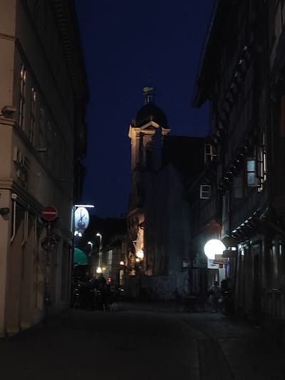 An alley in Göttingen, Germany, at night. The alley is bordered by buildings, one side featuring timber-framed construction. In the distance, a tower-like structure is visible under a dark blue sky. Illumination comes from streetlights and lights from buildings. People are gathered near the lights on the left side of the alley. The ground is paved. A "no entry" traffic sign is visible.
