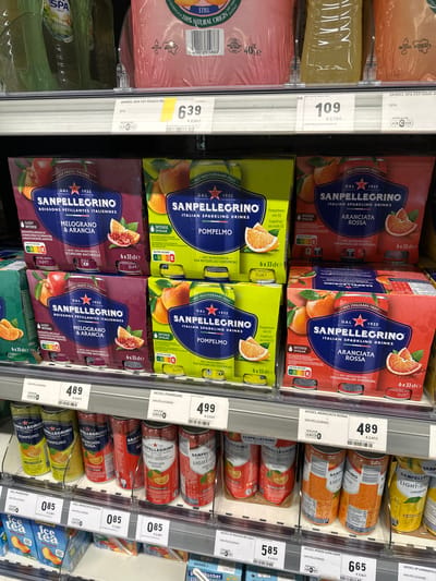 The image depicts a retail environment, likely a supermarket aisle, focusing on a display of Sanpellegrino sparkling drinks. The products are arranged on shelves, with several different flavors visible, including Melograno & Arancia, Pomplemo, and Aranciata Rossa. Some products are in multipacks of cans, while others are in larger packaging containing multiple cans. The lighting suggests an indoor setting, typical of a retail store. The image does not provide any specific visual cues that would indicate the location of Echternach, Luxembourg.