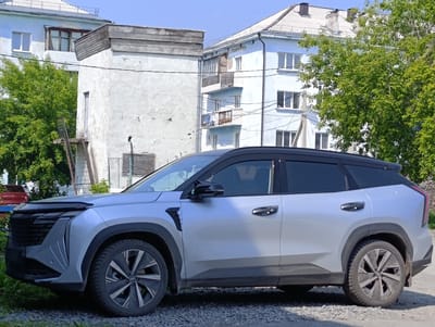 A silver SUV is parked on a gravel surface in front of two apartment buildings in Bogotol, Russia. The SUV is positioned in profile, facing right. The buildings appear to be aged, with light blue and off-white facades. Trees with green foliage are visible to the left and right of the car. The sky is clear and blue. The front of the SUV features a dark grille and silver accents. The side of the car shows black door handles and side mirrors. The rear of the vehicle is partially visible on the right.