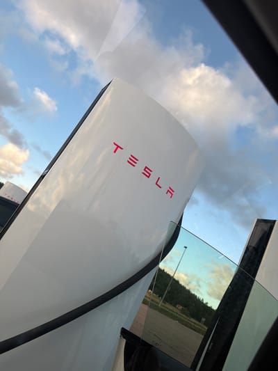 The image captures a close-up view of a Tesla Supercharger station, likely located in Hogstorp, Sweden, given the context. Two white Supercharger units are visible, with the prominent Tesla logo in bright pink lettering on the front of the closer unit. The sky above is filled with a mix of blue and white clouds, illuminated by what appears to be late afternoon or early evening sunlight.
In the foreground, a car window is present, reflecting the sky and a portion of the surrounding environment. Through the tinted glass, a landscape can be seen, featuring a paved road, trees forming a hillside in the distance, and a single lamppost. A small building is also discernible among the trees. The overall scene suggests an outdoor charging location, possibly near a rural or semi-rural area. The angle of the photograph, taken from within a vehicle, implies the photographer is a passenger or driver at the charging station. There are no people visible in the frame.