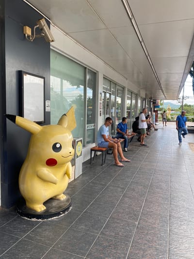The image shows an outdoor scene in Ban Ko Pru, Thailand. A large, yellow Pikachu statue lies on its side in the foreground. Behind it, a building has a security camera and a blank sign. A row of people sits on chairs along the building's walkway. Most appear to be middle-aged men. One man walks in the distance. The ground is tiled. The sky is visible in the background.