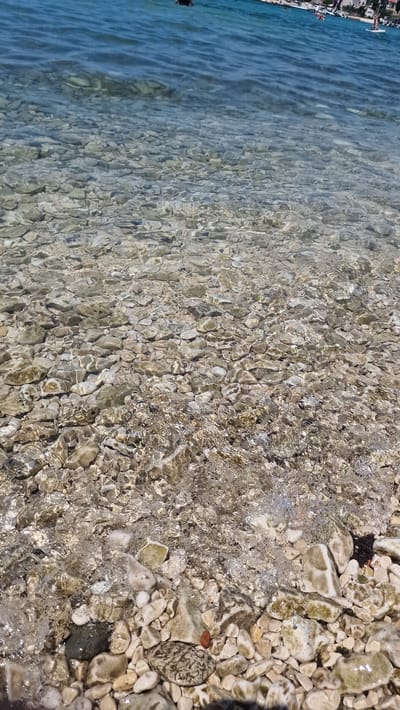 The image depicts a shallow, clear body of water with a pebble-strewn bottom. The water is a vibrant blue on the left side, gently transitioning to a more muted, transparent hue as it moves towards the right. Sunlight filters through the water, creating shimmering patterns and highlighting the varied colors and textures of the stones beneath. The pebbles range in color from light gray and white to earthy browns and greens, with some appearing smooth and rounded while others are more angular.

Along the left edge of the image, there's a glimpse of a shoreline with buildings and structures, suggesting a populated area. A few small figures, likely people, are visible near the water's edge, and one person appears to be in the water. The presence of buildings and the clear, inviting water suggest a beach or waterfront area, likely in a Mediterranean setting given the location context of Zadar, Croatia.

The time of day appears to be sunny, judging by the bright light and the reflections on the water's surface. The overall impression is one of a calm and serene day at the seaside, with the clear water and natural beauty of the pebbles creating a peaceful scene. There is no visible text in the image.