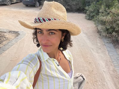 A young adult female is depicted in a close-up, selfie-style photograph taken outdoors in Aljezur, Portugal. She has curly dark brown hair, olive skin, and light-colored eyes, appearing to be green or hazel. She wears a wide-brimmed straw hat with a decorative band featuring red, dark green, and off-white stripes. Her attire consists of a light-colored, possibly white or cream, long-sleeved button-up shirt with thin yellow vertical stripes, worn open to reveal a lavender or light purple lace-patterned top or bralette underneath. She accessorizes with gold hoop earrings and a dark beaded necklace with a small, circular pendant. A light brown strap, likely from a bag, is visible over her right shoulder (viewer's left), and a folded light-colored garment, possibly a towel or sweater, is draped over her left arm (viewer's right). Her expression is a subtle, closed-mouth smile directed towards the camera.

The background reveals a dry, unpaved dirt or gravel pathway. To the immediate left, the path curves, and a square metal grate, presumably a drain cover, is visible on the ground. Adjacent to the path, a short, grey paved edge constructed from rectangular stones runs along portions of the ground. Further in the left background, two dark-colored parked vehicles, appearing to be sedans or hatchbacks, are partially visible. The landscape beyond the path on the right consists of dense, green and silvery-green shrubbery and low-lying trees, consistent with coastal or Mediterranean vegetation. The scene is illuminated by natural daylight, casting soft shadows.