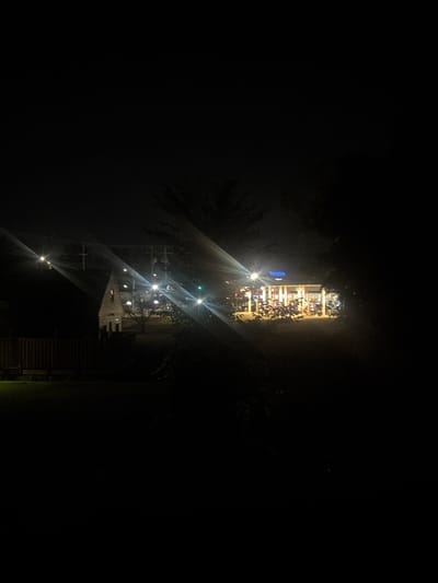The image depicts an outdoor scene at night, likely in a semi-urban or suburban area. In the middle ground, a building with a distinct A-frame roof and wooden siding is visible. The building is illuminated by nearby lights, which create prominent lens flares due to the camera's exposure to bright light sources. To the right of this building, a larger structure, possibly a business or gas station, is illuminated with a bright blue sign and multiple lights. This structure appears to have a covered area with what looks like gas pumps and several motorcycles parked in front. The foreground is dark with silhouettes of trees and foliage obscuring parts of the scene. A wooden fence is also visible in the lower left portion of the image. The overall impression is a night view of illuminated buildings amidst darkness, with some vegetation and a fence indicating a residential or commercial perimeter.