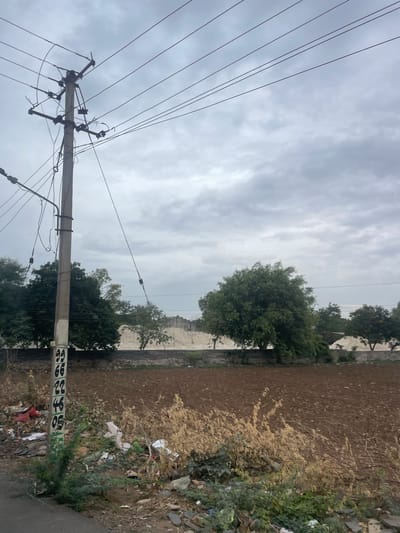 This image depicts an outdoor scene near Bethamcherla, India, featuring a utility pole with multiple electrical wires against a cloudy sky. The pole, located on the left side of the frame, has markings that appear to be numbers, with "99", "66", "22", "46", and "05" visible. The pole is surrounded by a significant amount of litter and debris on the ground. Behind the pole, there is a field of tilled brown soil, bordered by a low stone wall and trees. Further in the background, there are piles of light-colored material, possibly construction or industrial waste, and more trees and vegetation. The overall environment suggests a somewhat undeveloped or transitional area within the city.