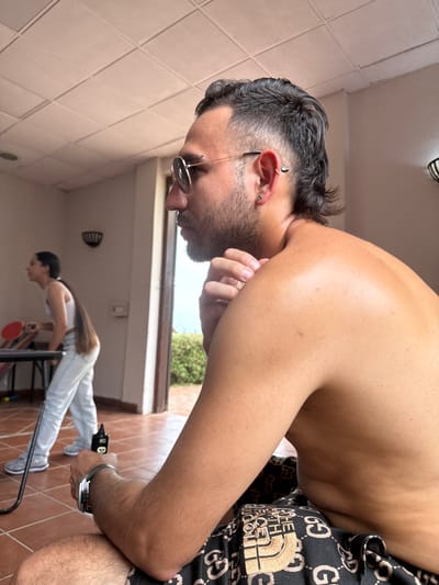 A man with a mullet hairstyle, earrings, and sunglasses stands close to the camera. He's shirtless, wearing shorts with a Gucci and North Face pattern. In the background, a person in light jeans and a white top is performing a plank exercise on a metal frame. A tiled wall and a glimpse of greenery are also visible.